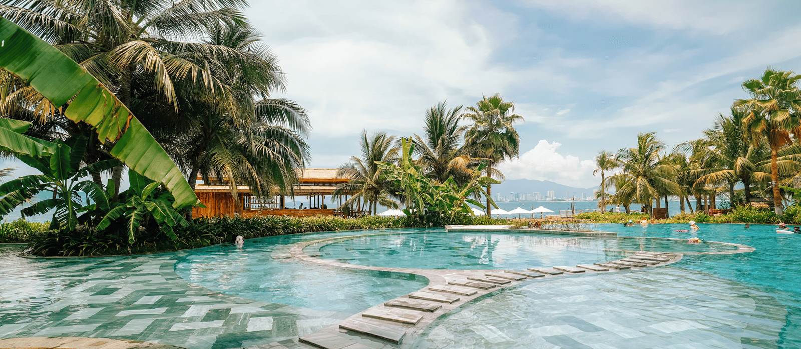 Swimming pool surrounded by palm trees at Alibu Resort in Vietnam, featuring several layers and narrow pathways in it where people can walk and go to different sections of the pool.