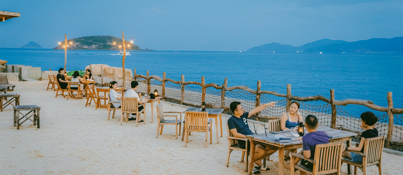 A seating area with table and chairs on the beach featuring people sitting on the chairs and enjoying the views of the sea.