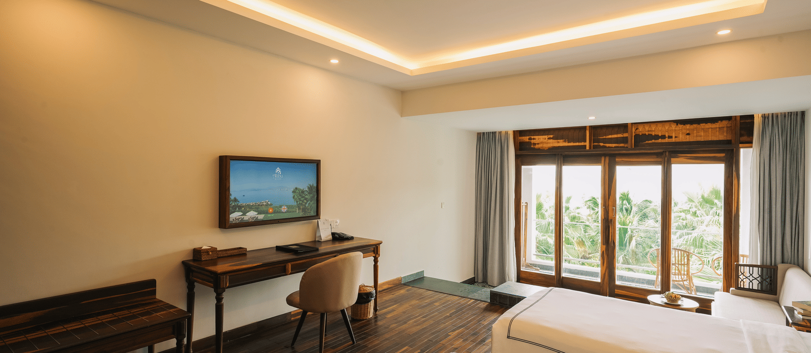 A bed at Alibu Resort Nha Trang featuring twin bed, a TV attached on the wall, a table, and windows with curtains.