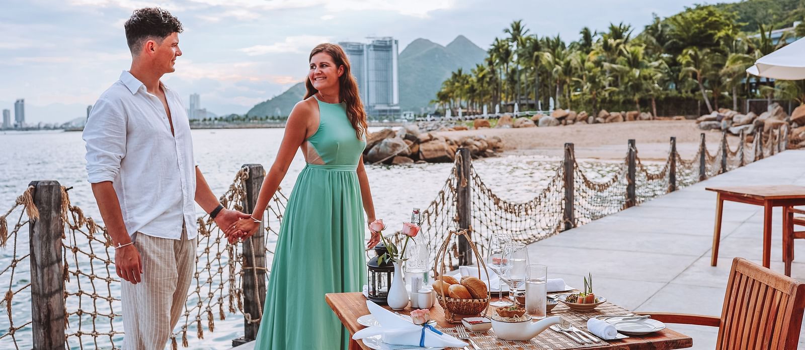 A couple hand in hand by the bay during daytime with views of the ocean, city and hills in the background - Alibu Resort Nha Trang