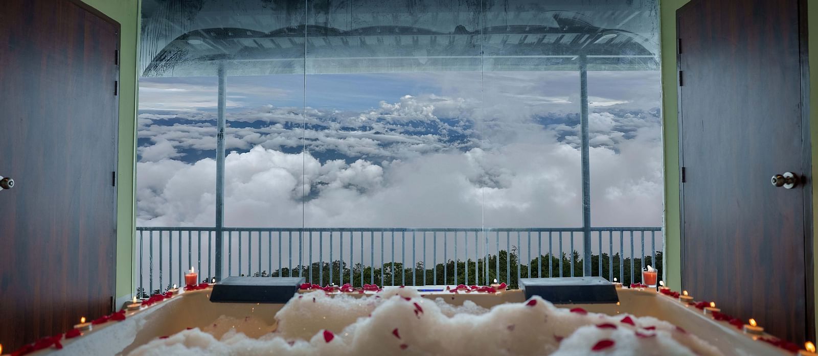 A jacuzzi with rose petals scattered, leading to a balcony overlooking a cloudy sky and distant hills, suggesting a romantic setup at Summit Enigma Resort and Spa.