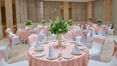 An event hall setup at KPG Sarovar Portico, Raj Nagar Extension, Ghaziabad featuring round dining tables, chairs with bows, flower vases, and wall panels.