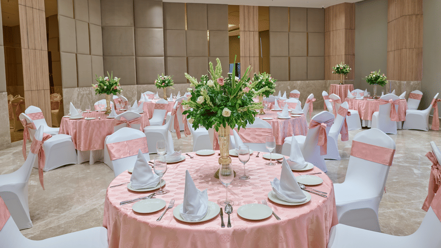An event hall setup at KPG Sarovar Portico, Raj Nagar Extension, Ghaziabad featuring round dining tables, chairs with bows, flower vases, and wall panels.