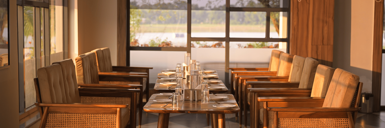 An elegant indoor seating area with wooden chairs and large windows framing outdoor garden views at Hotel Sonar Bangla Mayapur.