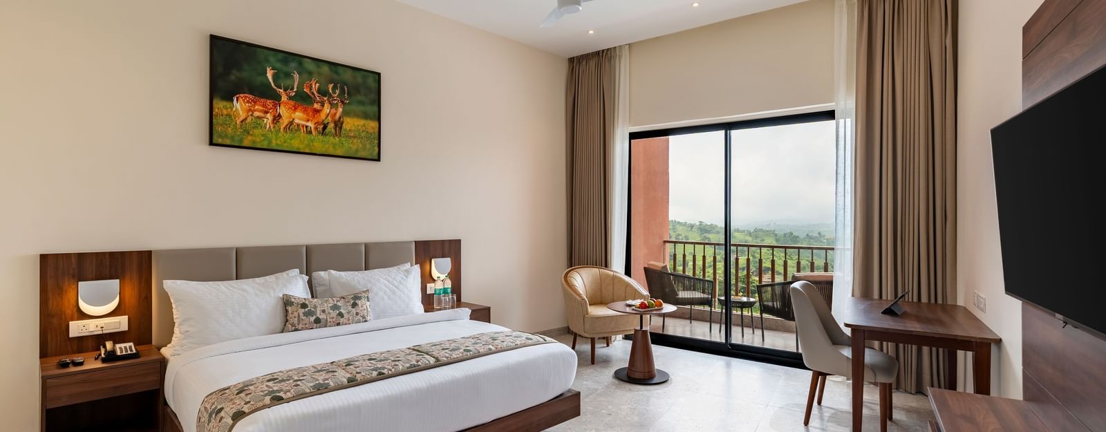 A spacious bedroom at Osian Valley Resort & Spa with a large bed, a desk and chair, a wall-mounted television, and a balcony with a scenic view.