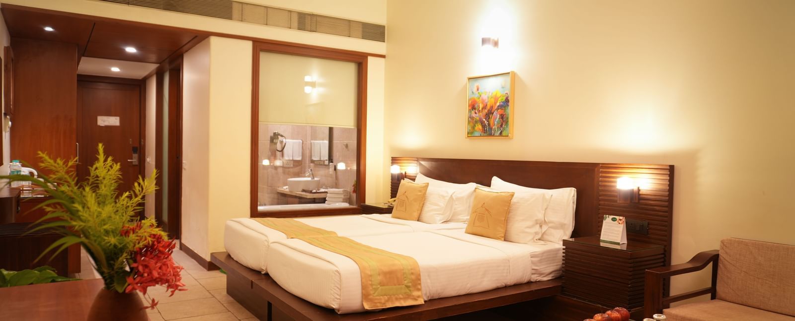 Standard Suite at Vedic Village Spa Resort showcasing a double bed with wooden headrest, tiled flooring, glass-panelled wall, glass-topped table, and a lounge couch.