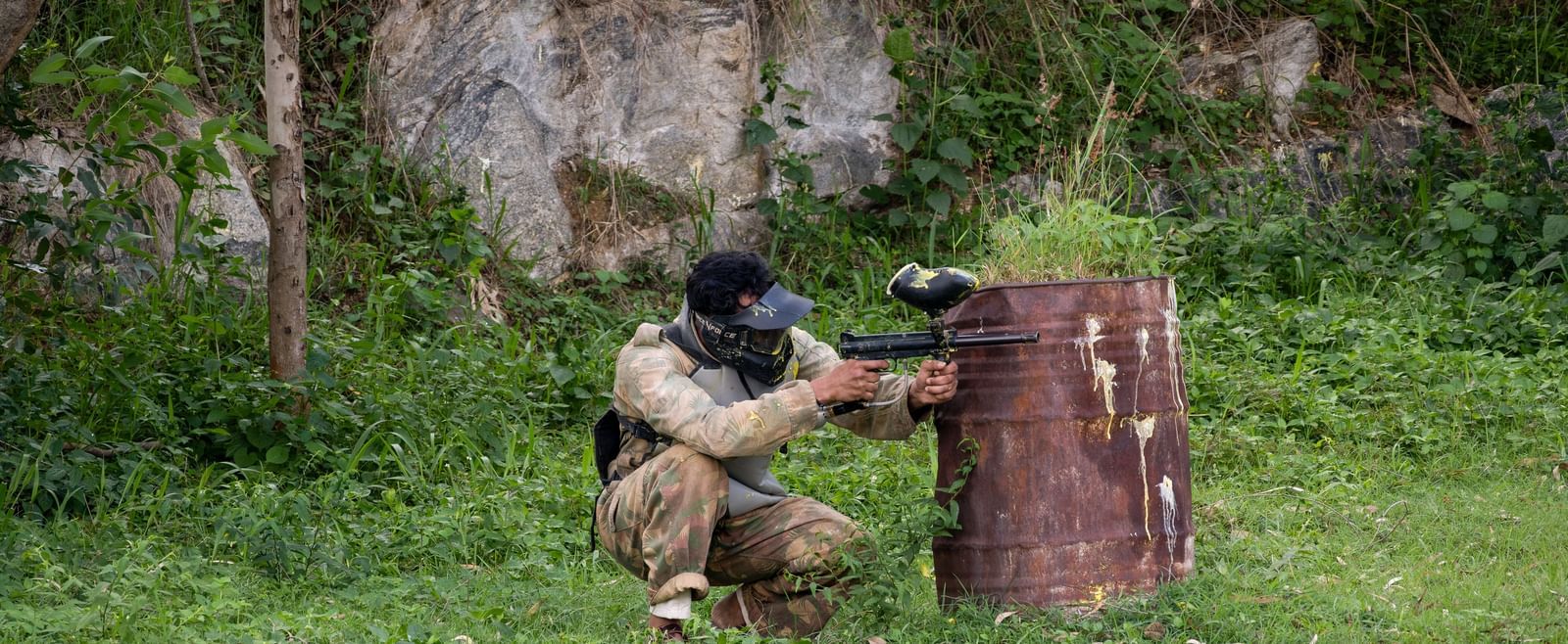 Two individuals are engaged in a paintball game in a natural setting, with one person taking cover behind a wooden barrier | RD's Nature Retreat, Bangalore