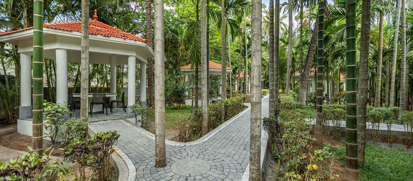 A white gazebo with an orange roof and a paved pathway winding through palm trees and greenery - Mango Hill Resort, Coimbatore