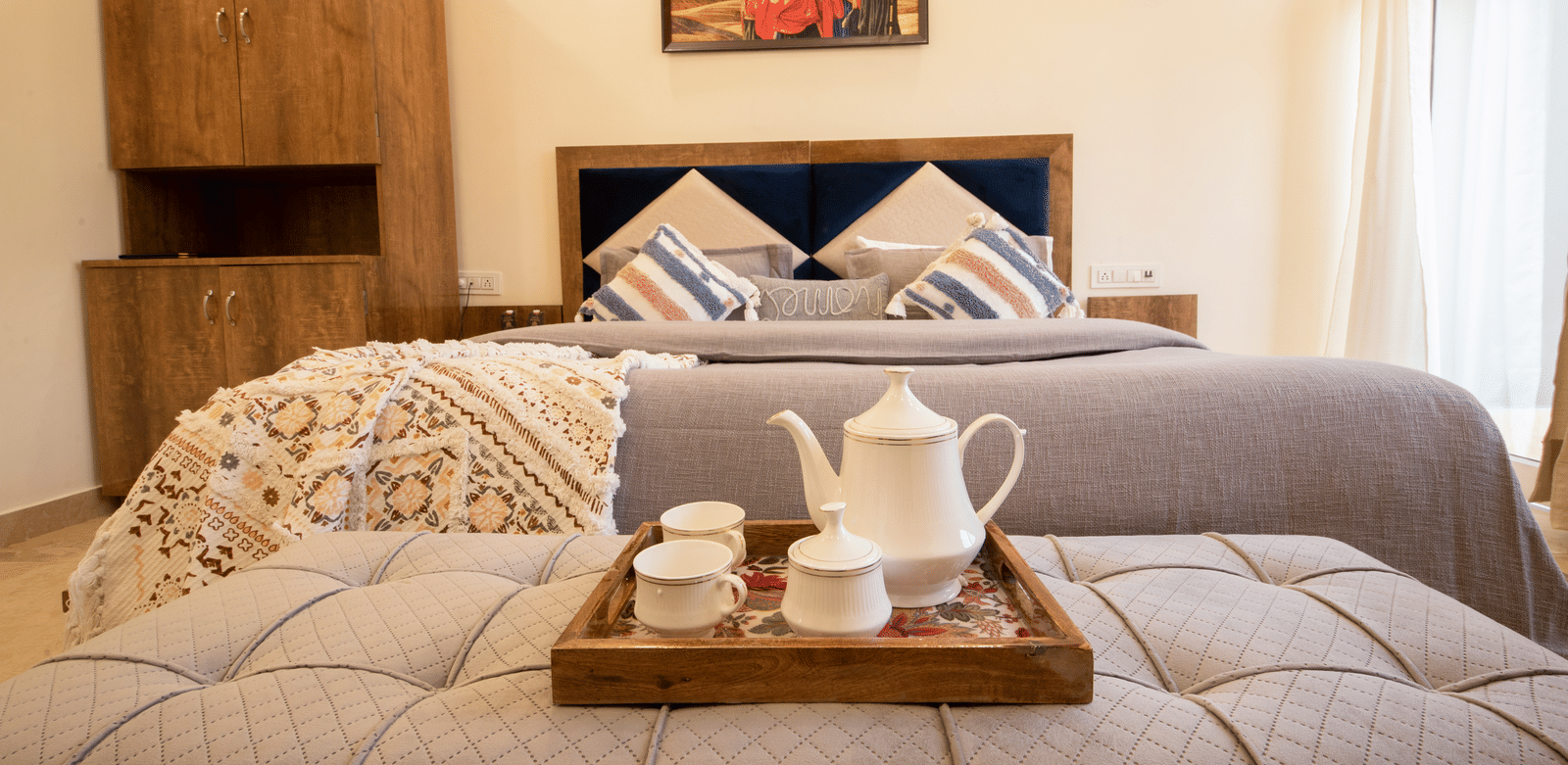 A hotel room, featuring a large grey bed with white linens, blue and orange pillows, a wooden tray with a teapot and cups on the bed, and a painting on the wall | Perfectstayz Value Hills, Rishikesh