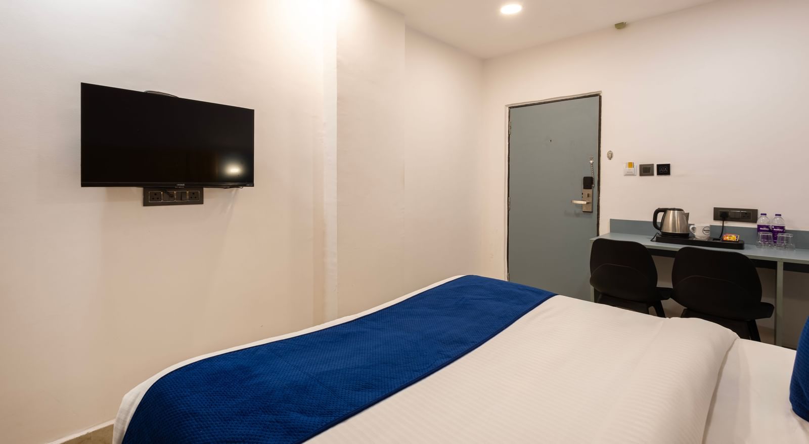 A Deluxe Room with a bed, a wall-mounted television, and a desk area with a chair and what appears to be a mini-fridge or coffee station. - Beestay Hotels Financial District, Hyderabad 