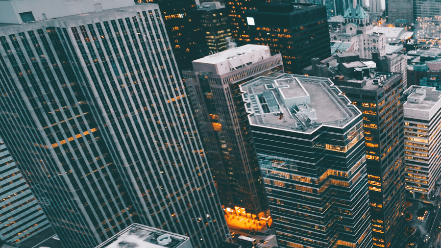 Aerial view of a dense city skyline at dusk with illuminated skyscrapers, rooftop details, busy streets, and a body of water in the distance.