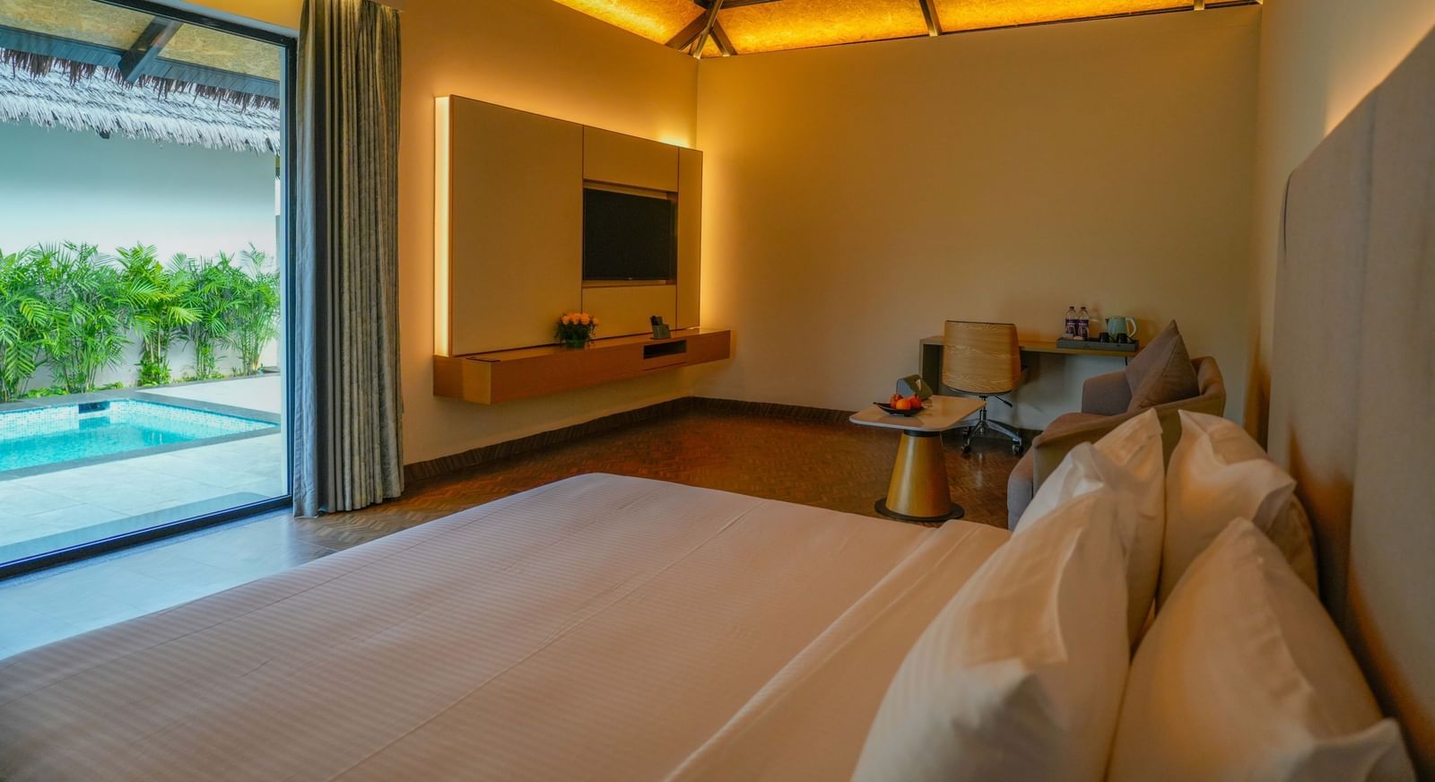 Interior view of a luxury villa room with a neatly made bed, warm ambient lighting, a mounted TV, a sitting area with a desk and chair, and a view of a private pool through the open sliding glass door - The Shore Trishvam, Pondicherry