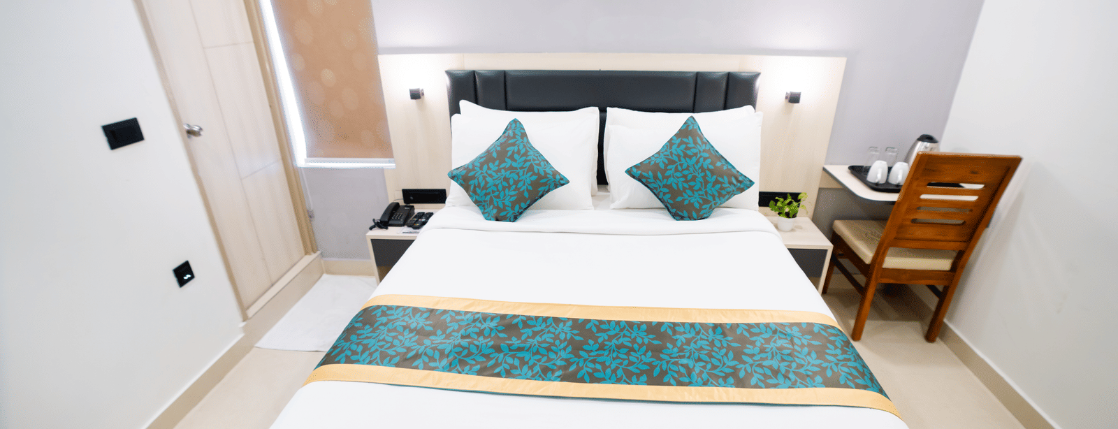 View of a room with a spacious bed, bedside table, wall-mounted A/C above the headboard, and a closed door to the side at Sunrise Business Hotel, Hyderabad.