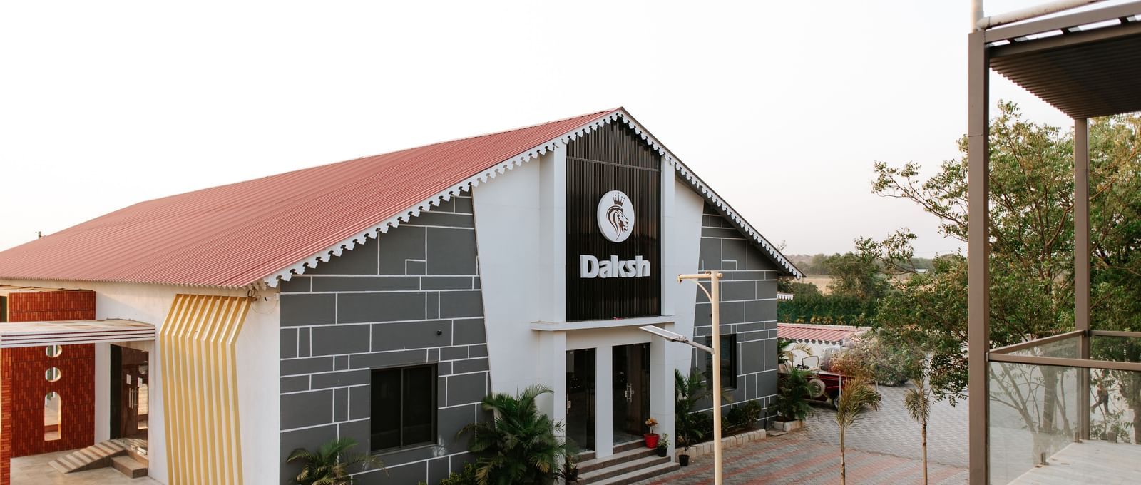 A side angle shot of a the exterior of Daksh Eden Greenz, Sasan Gir on a bright day, surrounded by trees and spacious walkways.