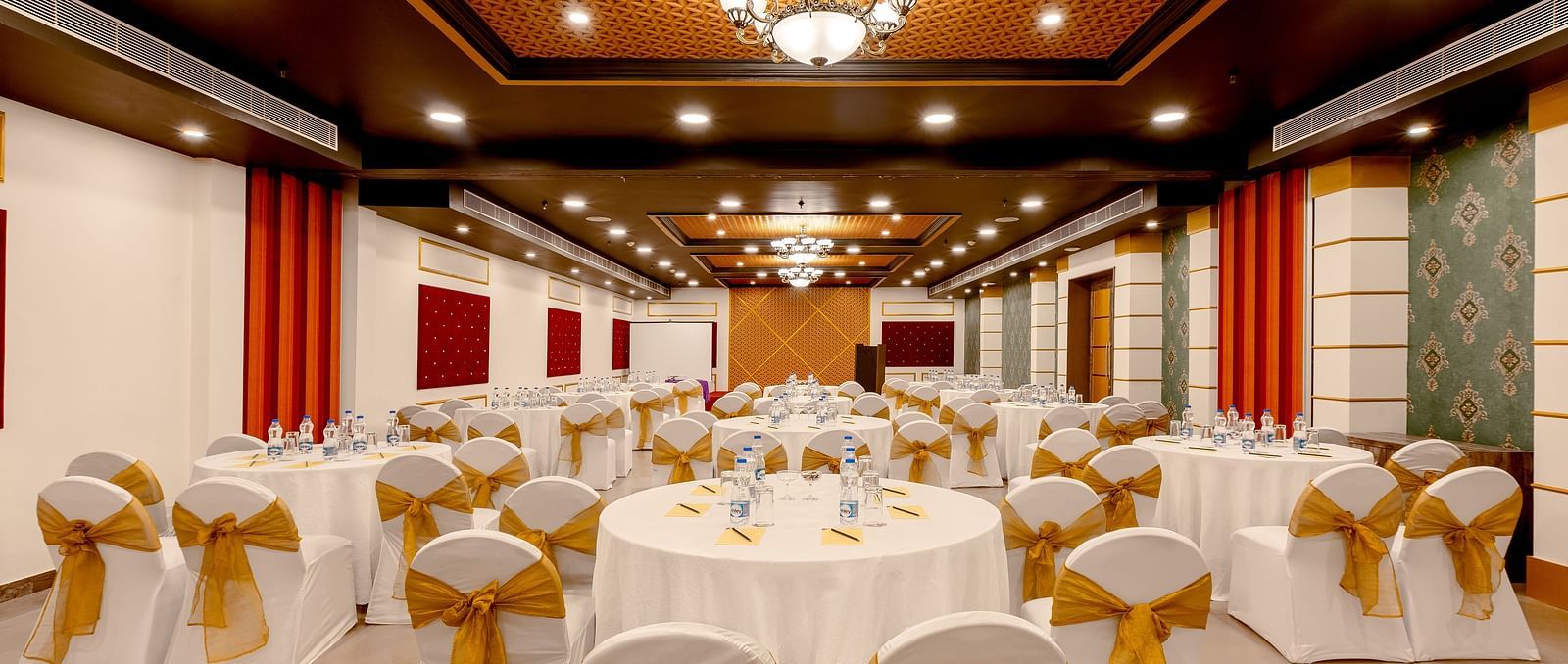A spacious banquet hall with round tables covered in white cloths and gold chair covers. The hall has a modern design with dark wood accents and a crystal chandelier.