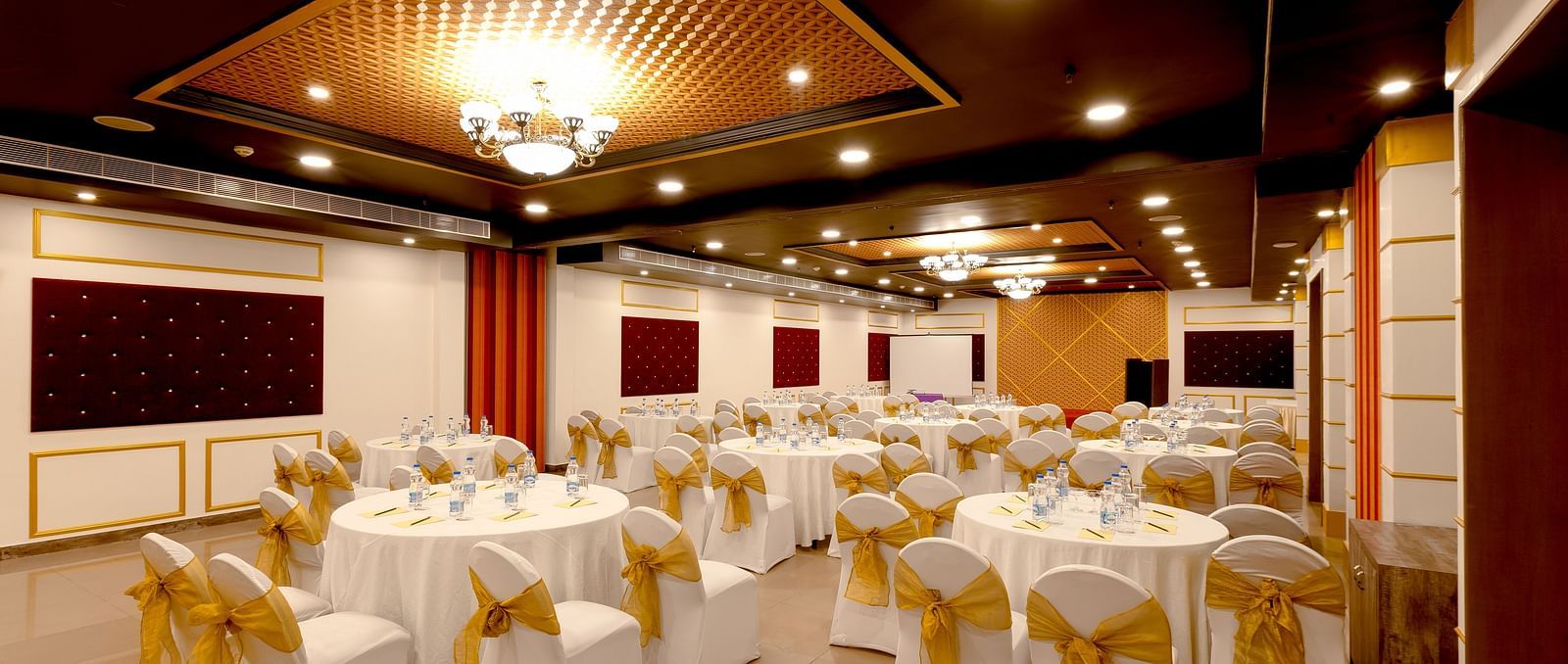 A spacious banquet hall with round tables covered in white cloths and gold chair covers. The hall has a modern design with dark wood accents and a crystal chandelier.