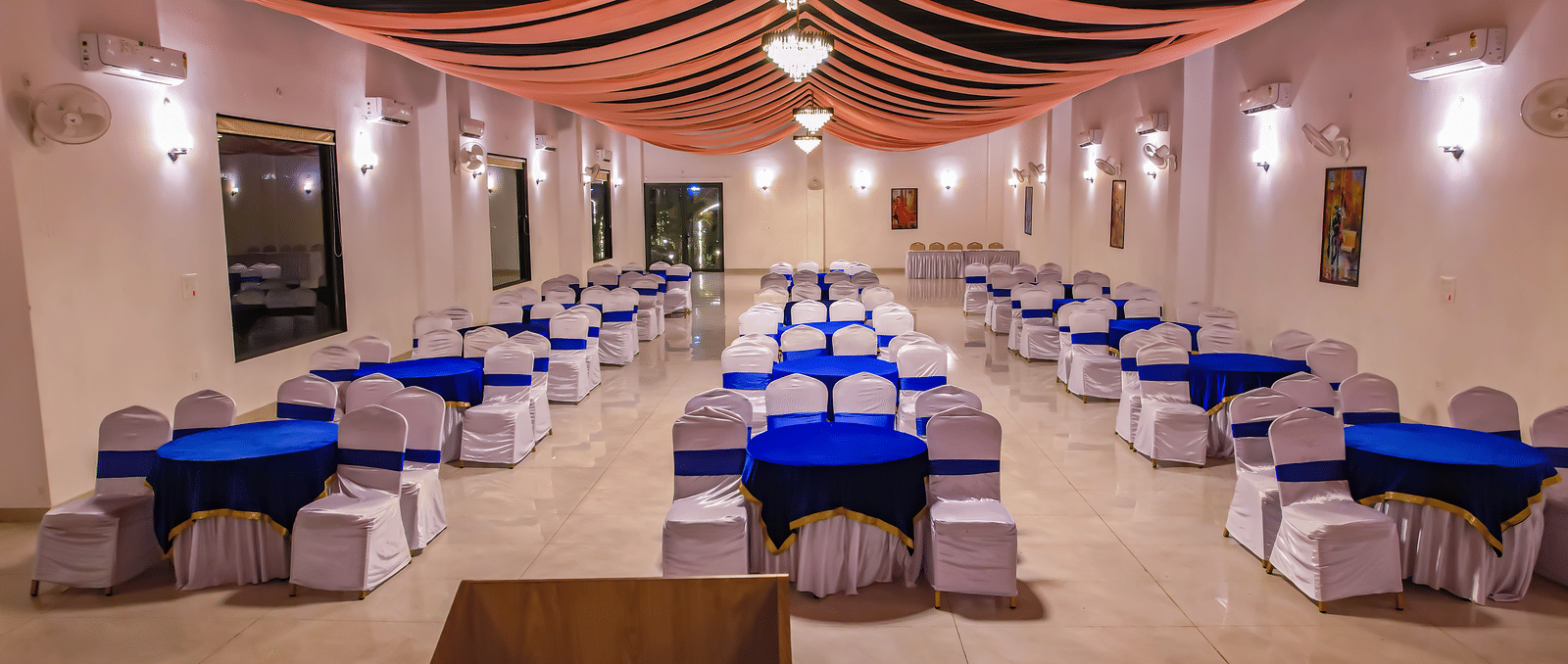 A long indoor dining hall at Vedikant Resorts The Mallard Corbett shows rows of tables and chairs beneath a decorated ceiling.