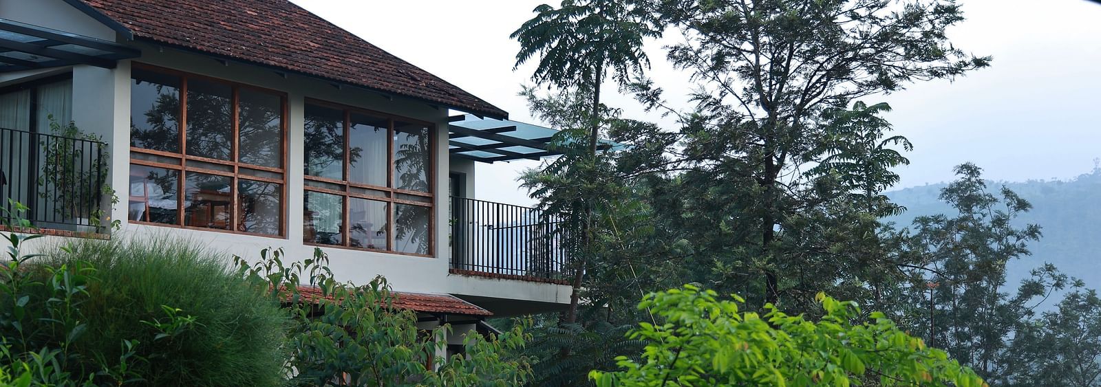  A building at Vaastha Vagamon, nestled amidst abundant foliage, with large windows overlooking a verdant landscape.