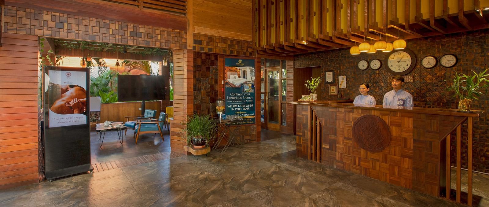 A spacious reception area featuring wooden accents with two people standing behind a wooden desk offering a seamless blend of rustic and modern elements. Large entrance letting in natural light, and the setting is calm and inviting - Coral Reef Resort & Sp