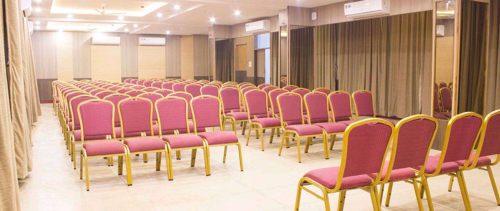 A wide shot of a banquet hall with multiple rows of empty pink and gold chairs. | Banquet Hall | Daksh The Madhuvan Suites