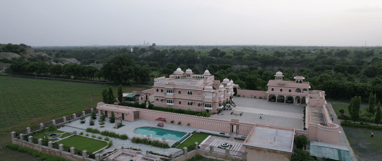 A high-angle aerial view of Mahendra Niwas, presenting the entire property with its intricate garden design, swimming pool, and surrounding landscape, all under a clear sky.