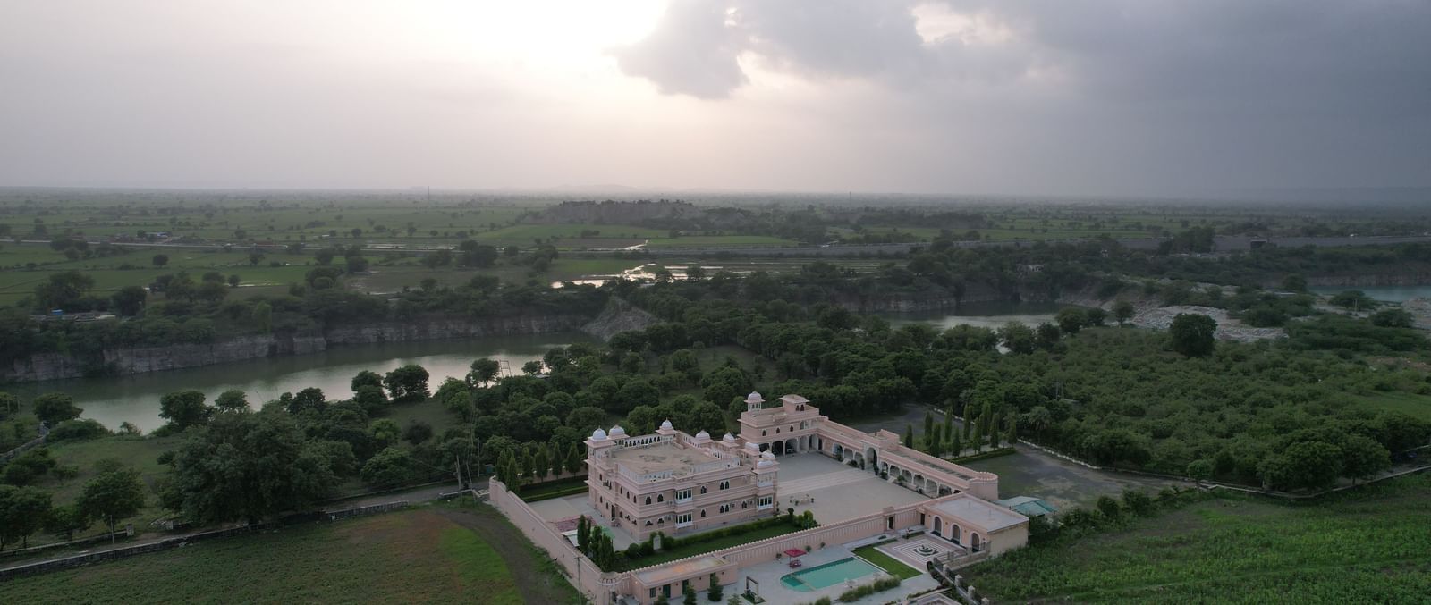 An expansive aerial view of Mahendra Niwas under a dramatic, cloudy sky, showcasing the property's grand scale and lush surroundings.