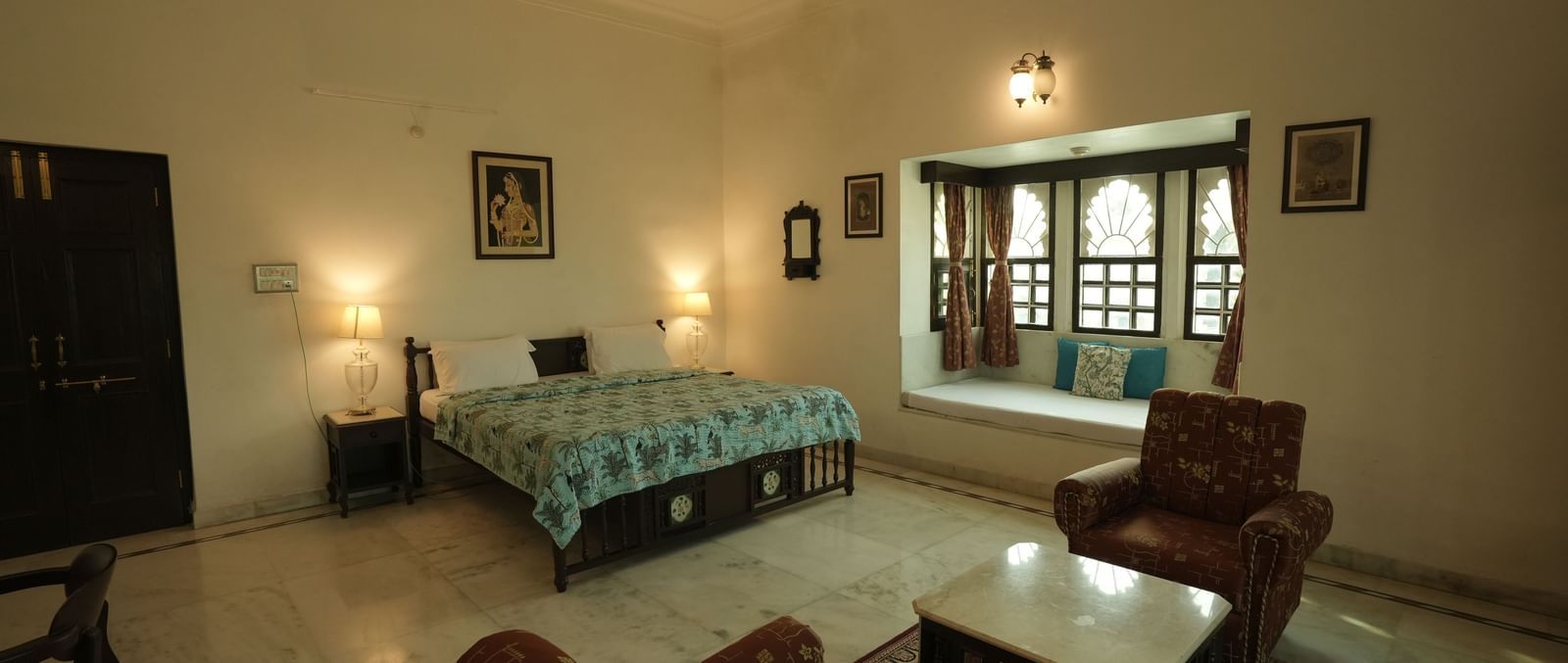 An eye-level shot of a bedroom with a large bed in the center. The deluxe room at Mahendra Niwas features a bay window seat and a pair of chairs around a low table.