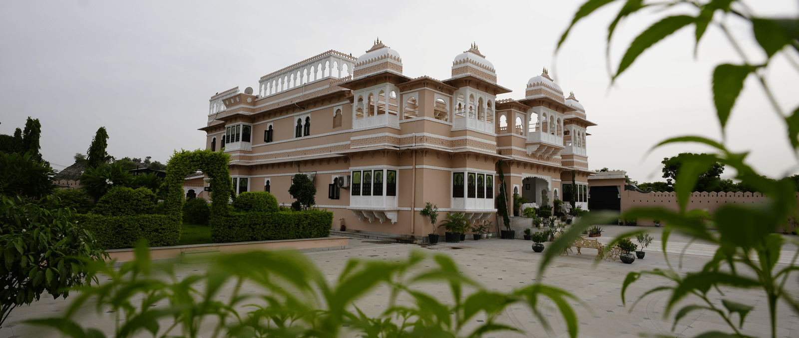 A low-angle exterior view of Mahendra Niwas, framed by vibrant green foliage, giving a sense of the property's elegant architecture nestled within a beautiful garden.