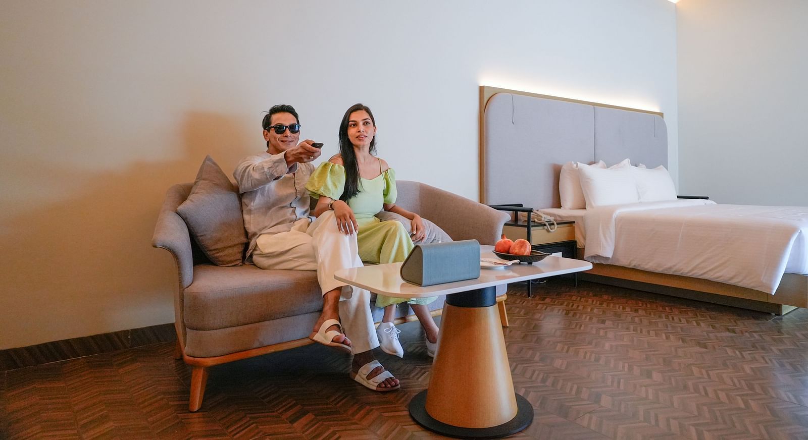 Couple relaxing on a sofa in a modern hotel room with a bed, coffee table, and warm lighting - The Shore Trishvam, Pondicherry