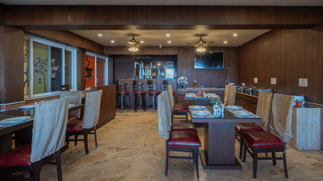 An indoor dining area at Perfectstayz Premium The Pride, Mussoorie with dining tables, chairs, counter space, and a wall-mounted television.