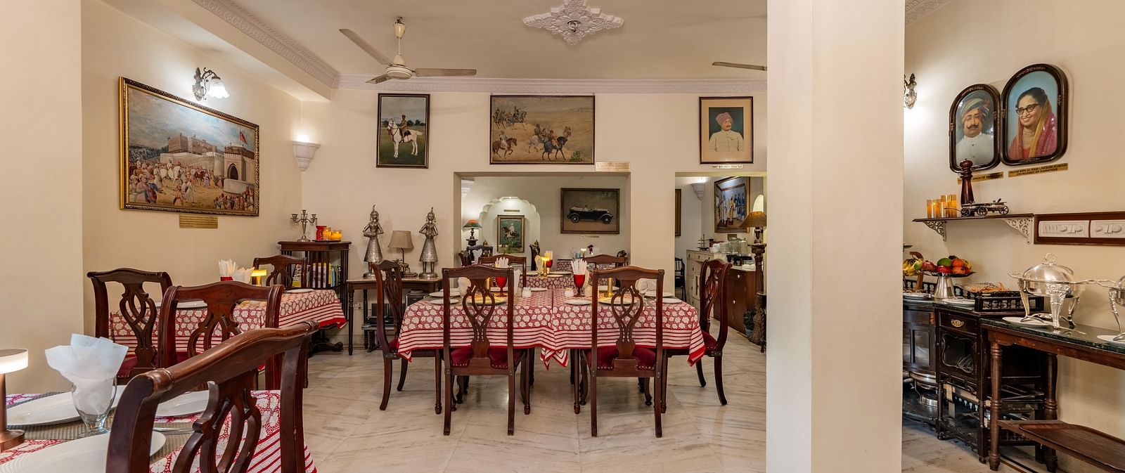 The inviting dining space at Dera Rawatsar, Jaipur, features multiple set tables with patterned cloths, wooden chairs, and an array of framed pictures on the walls.