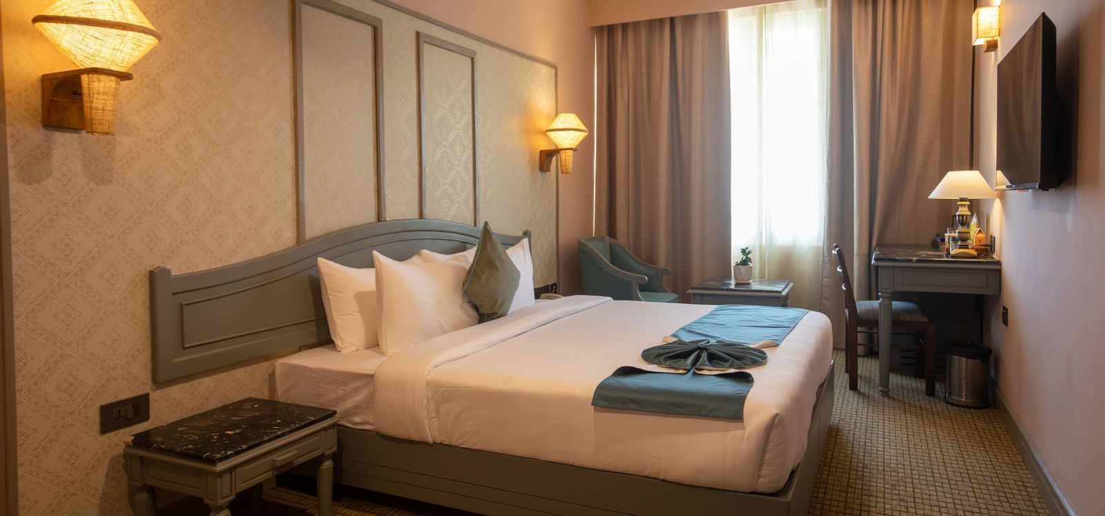 A guest room at Shilton International, Kochi, featuring a comfortable bed, a workspace, and elegant wall sconces.