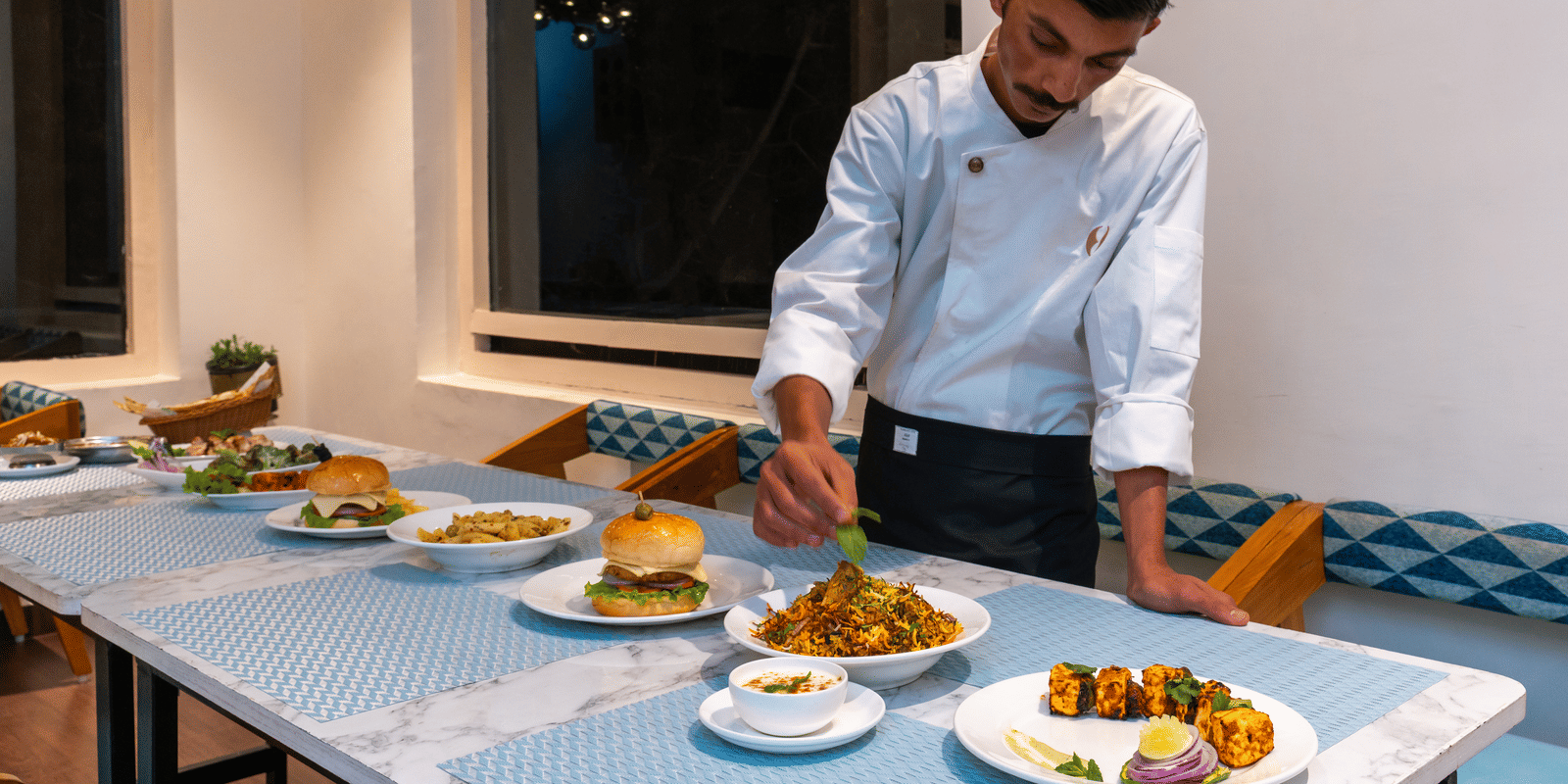 A chef in white uniform carefully plating a row of gourmet burgers in a modern kitchen at Himalayas Resort By The Lake Hill, Mukteshwar.