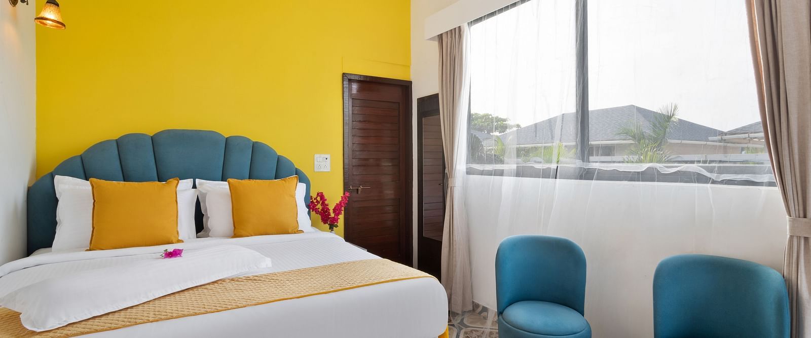 A bright, modern bedroom, a large window, a cosy double bed, and teal armchairs.