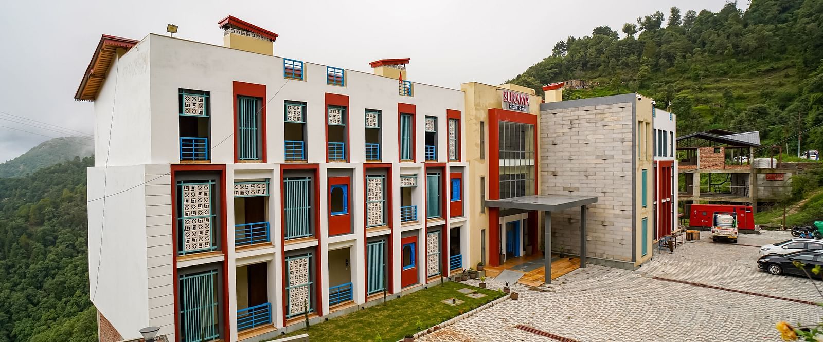 An aerial view shows a multi-story building complex with red and white facades situated on a hillside | Sukama Resort & Spa, Nainital