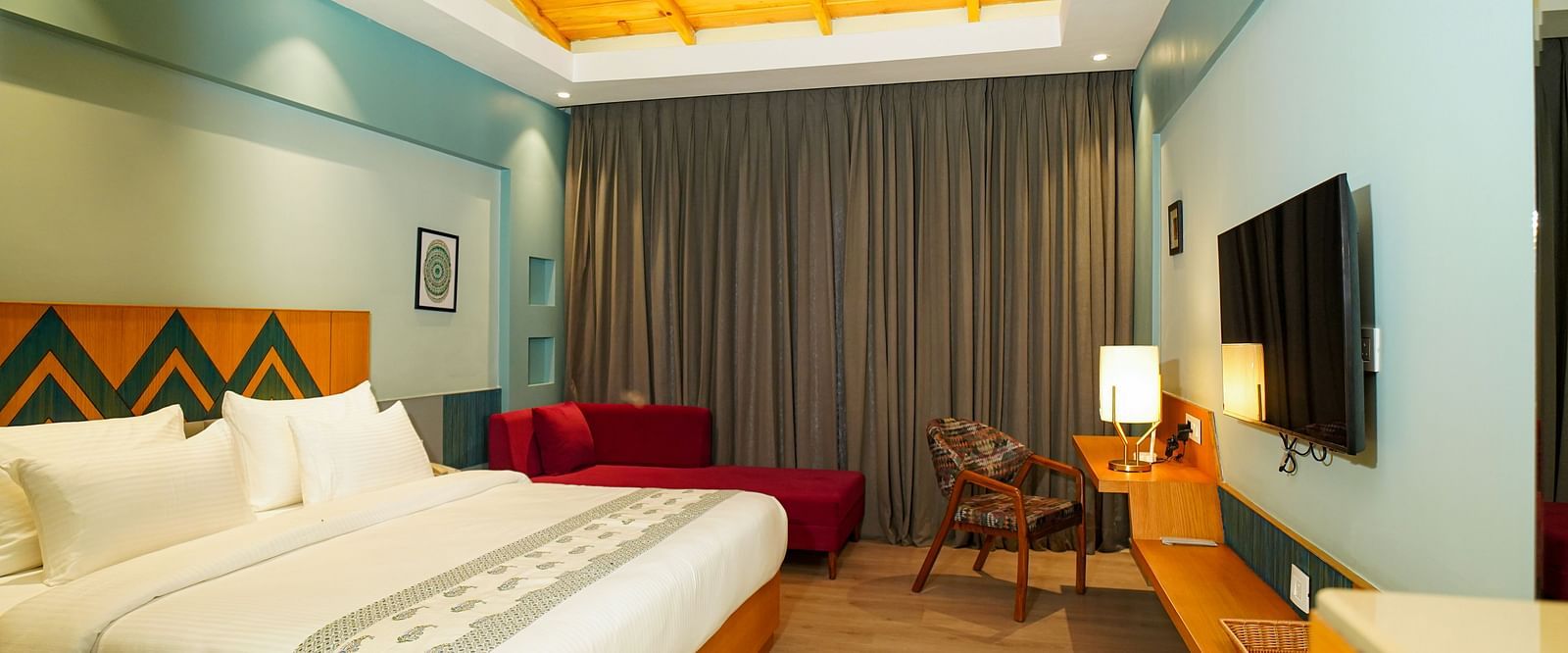 A hotel room with a large bed featuring white linens and a wooden headboard also includes a wall-mounted television and a view of curtains and a patterned wall | Deluxe Room with Valley View | Sukama Resort & Spa, Nainital