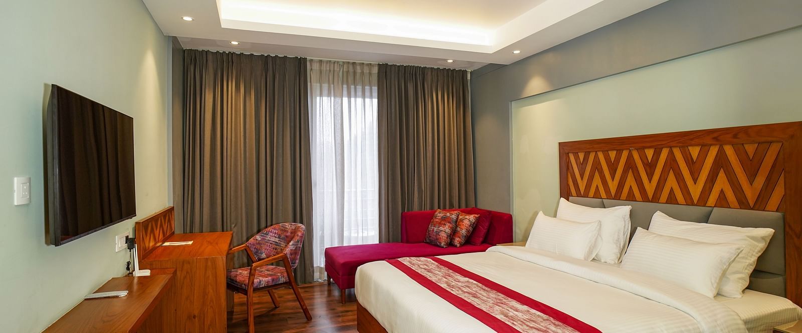 A hotel bedroom shows a neatly made bed with red accents, a sofa, and a television on the opposite wall | Deluxe Room with Himalayan View | Sukama Resort & Spa, Nainital