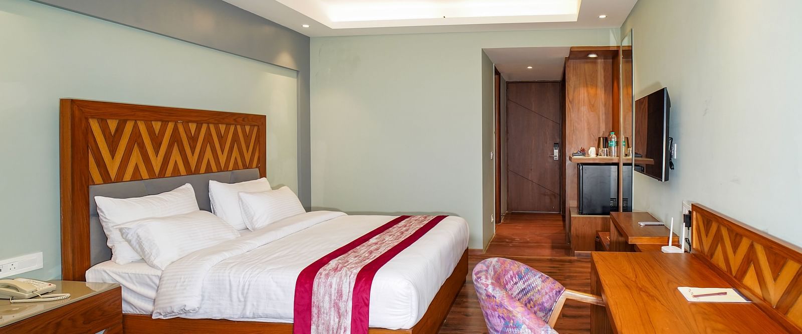 A spacious hotel bedroom is furnished with a large bed, a desk area, and a television, all in warm tones | Deluxe Room with Himalayan View | Sukama Resort & Spa, Nainital