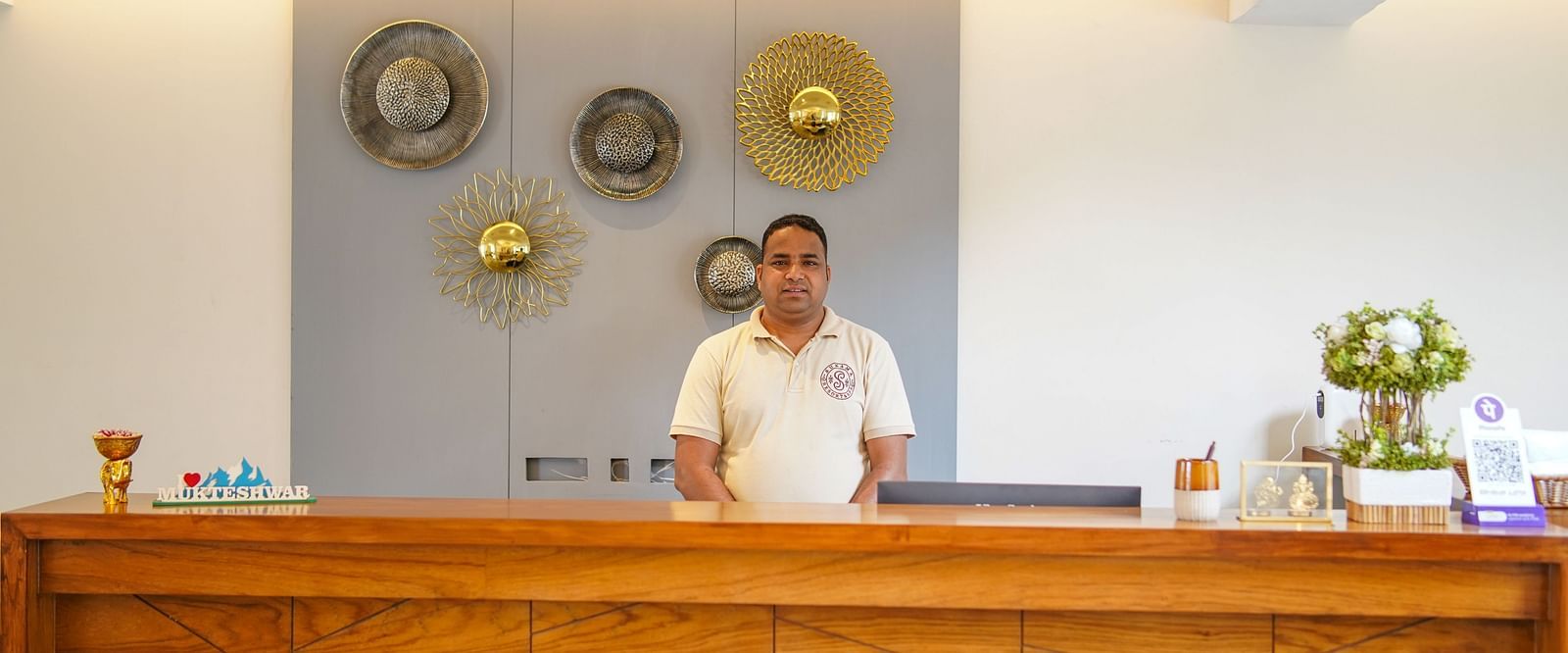 A hotel reception desk with a person standing behind it, and decorative plates on the wall behind them | Sukama Resort & Spa, Nainital
