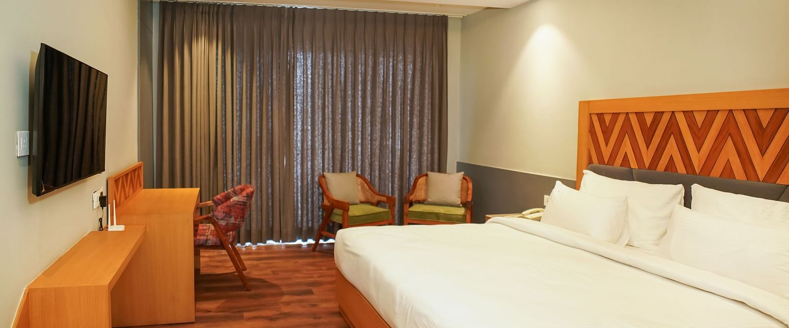 A modern hotel room with a large bed, wooden headboard, and seating area, lit by warm overhead lighting | Deluxe Room | Sukama Resort & Spa, Nainital