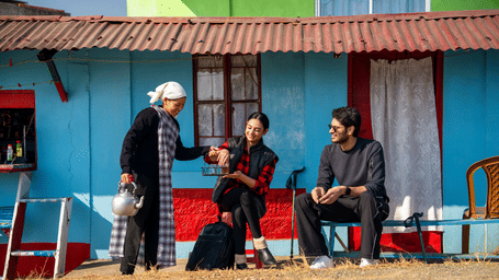 Guests enjoy tea served by a local Khasi woman pouring from a kettle outside a blue Cherrapunji village house with a red tin roof and lace curtains.
