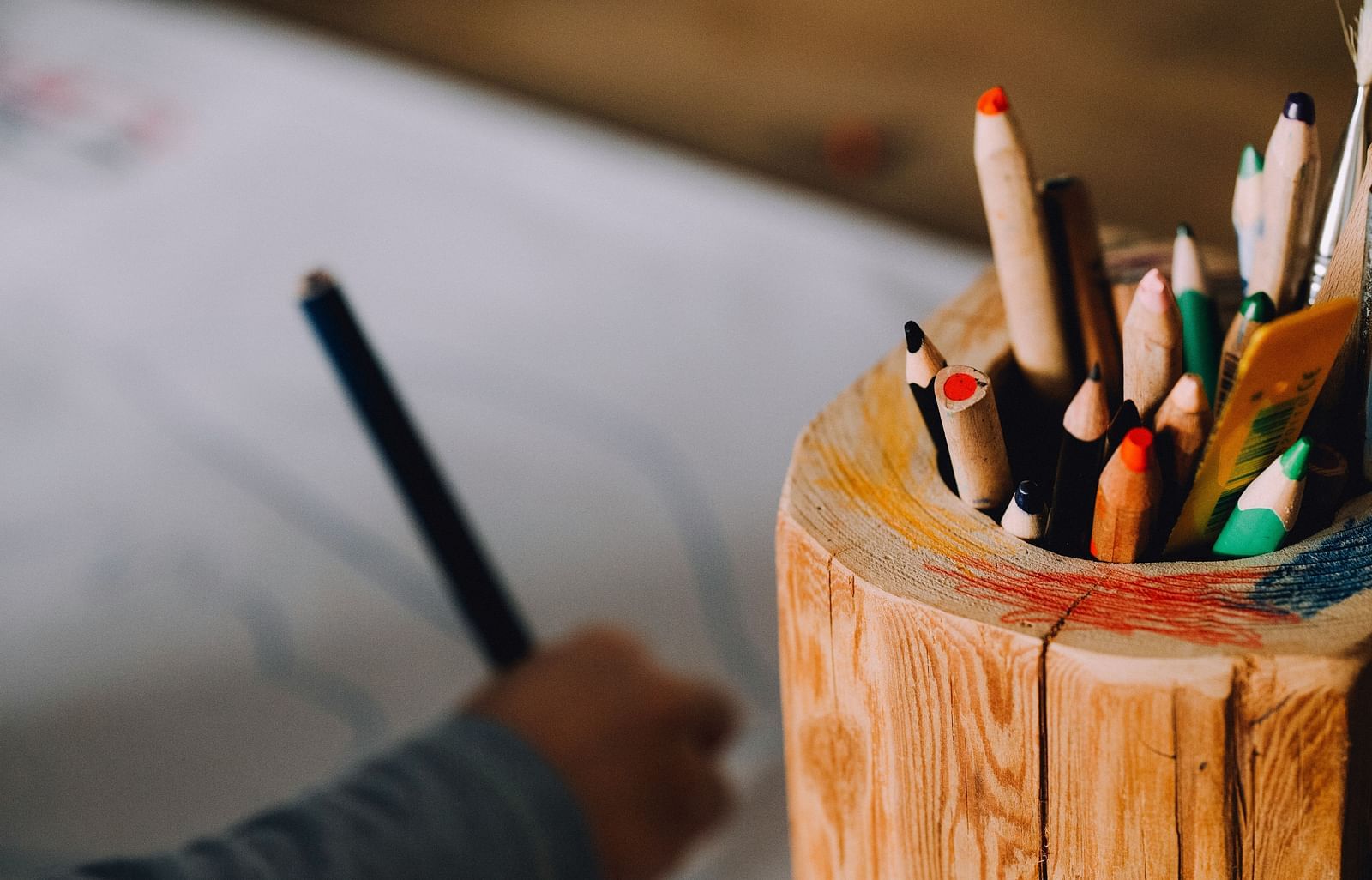 A wooden box overflowing with colorful pencils, while a child's hand in the background delicately sketches on paper.