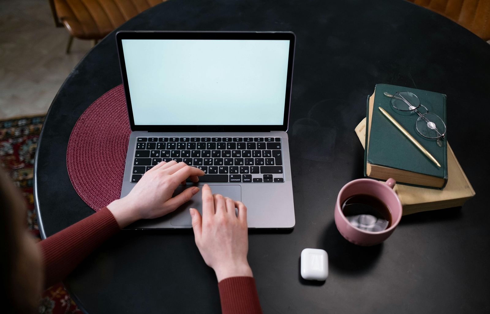 A person is using a laptop placed on a black table. To the right of the laptop, there’s a coffee mug and next to it are spectacles and a diary.