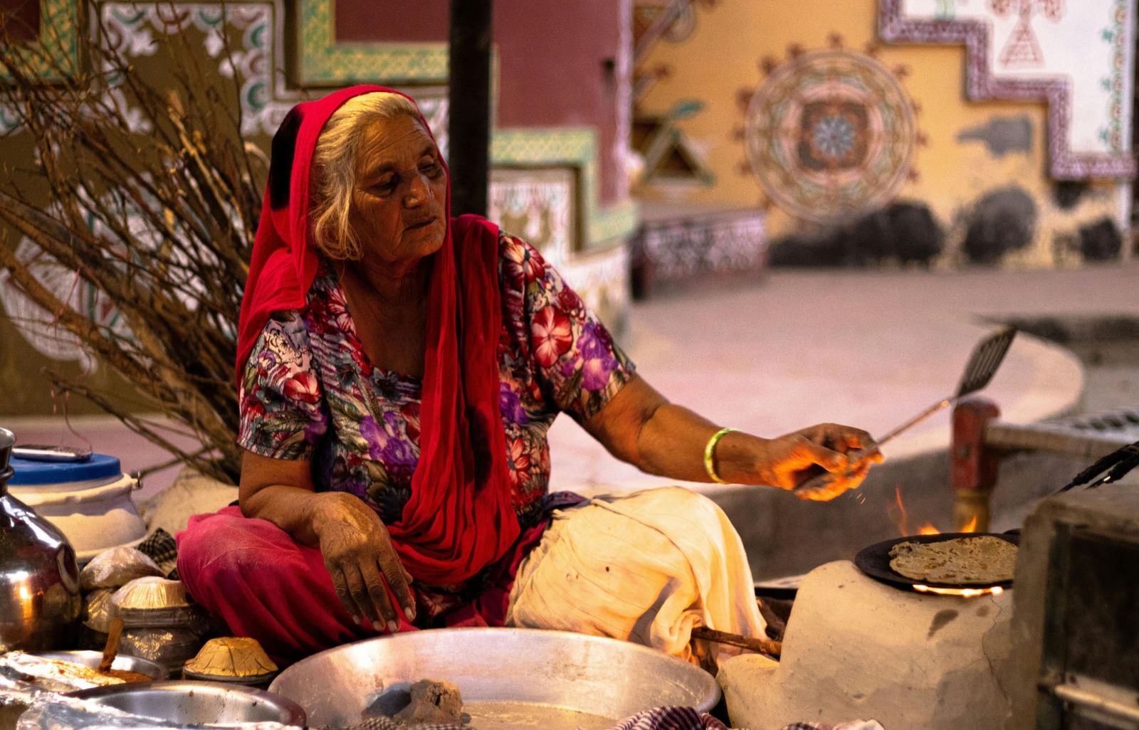 A woman in traditional attire is cooking over a fire outdoors, possibly part of an experience offered at Royal Heritage Haveli.