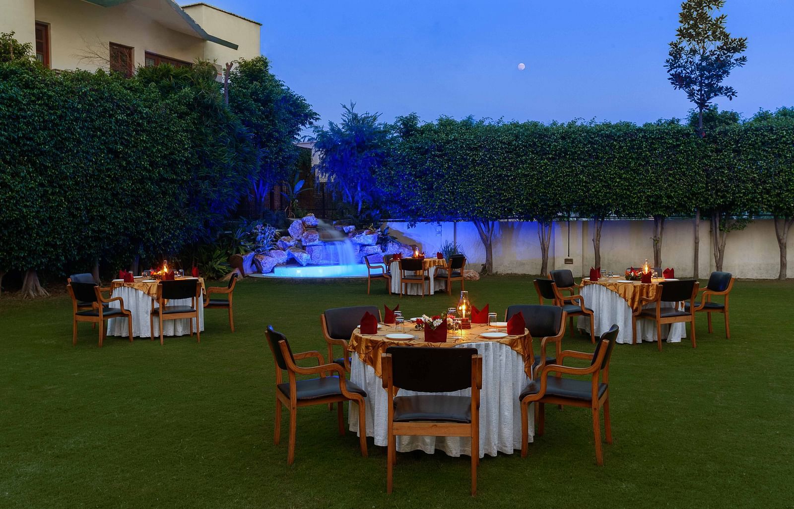 An open lawn with tables and chairs arranged in a circle, each adorned with decorative settings and a small lamp, featuring a lit-up fountain in the corner