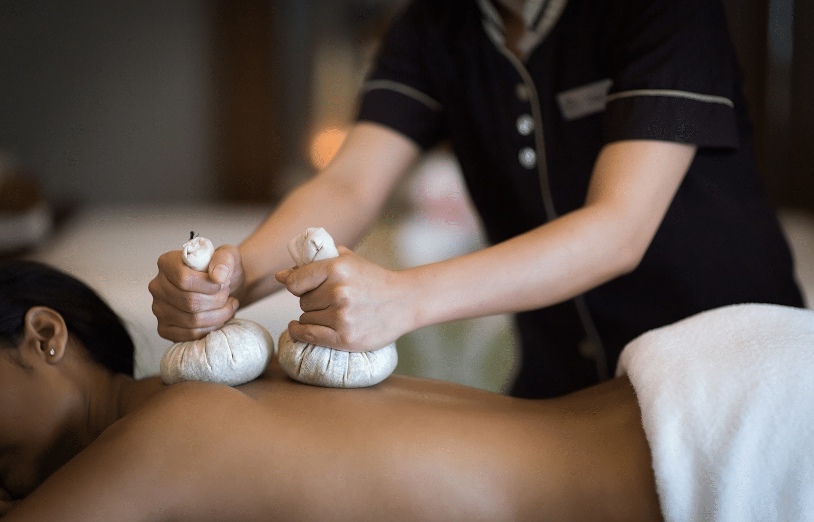 A masseuse massaging the client wearing a white towel, with two white massage kit on their back