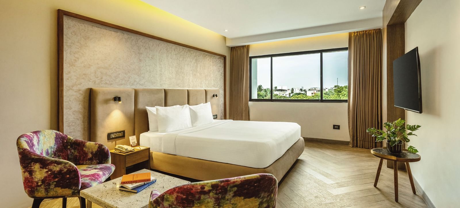 A wide-angle view of a room with a large bed at Mango Hill Central, Chennai. There are a chair and a small table in the foreground.