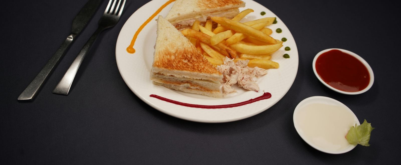 A plate of sandwich, is served with dipping sauces and a lime wedge on a dark surface.