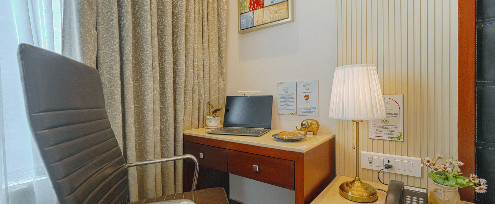 A close-up of a wooden hotel desk with a laptop, lamp, stationary, and a brown leather chair at BluSalzz Select City Centre, Amritsar.