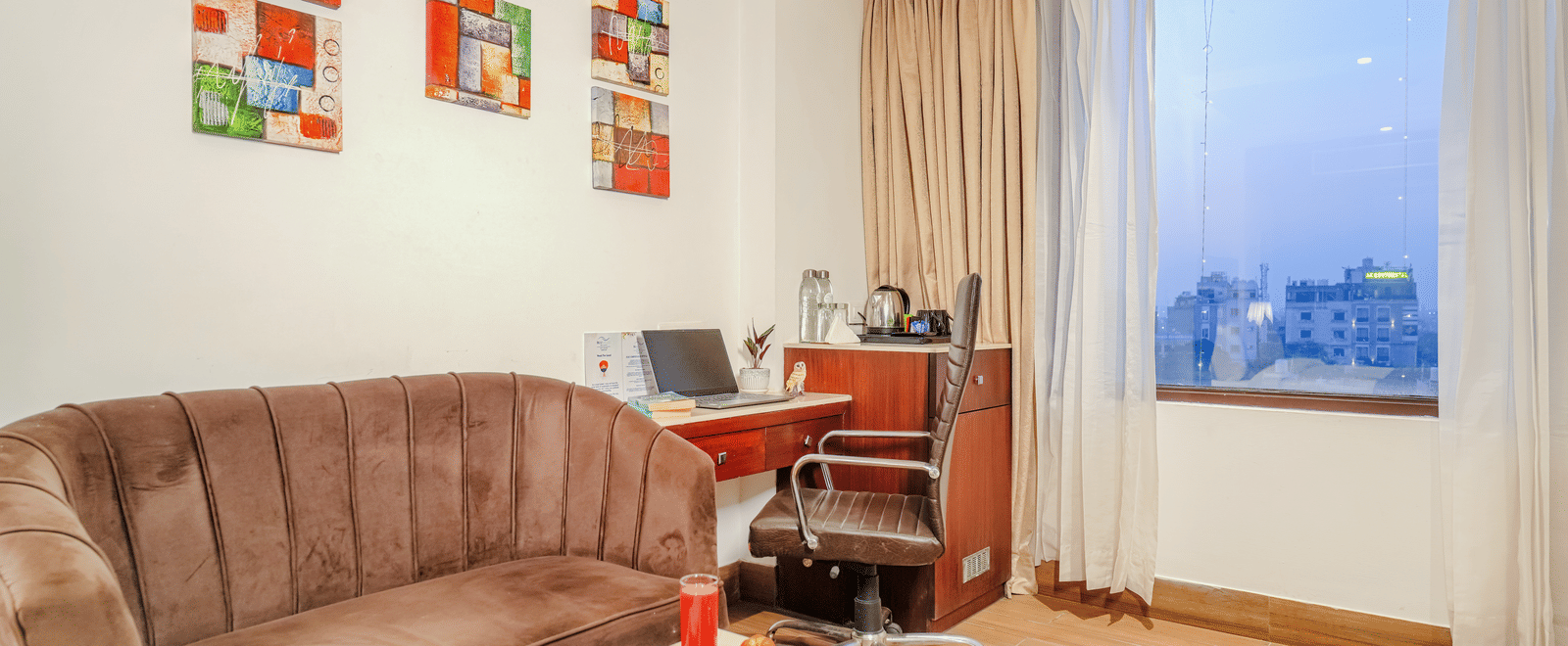 A small sitting and working area in a hotel room with a brown velvet sofa, a laptop on a desk, and colourful wall art at BluSalzz Select City Centre, Amritsar.